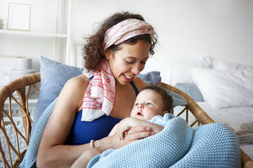 portrait of cheerful joyful young hispanic mother relaxing in weaven chair at home, talking to her b