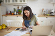 © shurkin_son - Indoor shot of attractive young female doing calculations, managing home budget, sitting at kitchen table and smiling. Pretty girl paying domestic bills online, using laptop computer and calculator