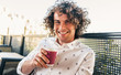 © Z A C - Horizontal closeup portrait of male with toothy smile, curly hair, wearing casual trendy shirt drinking a cup of coffee in the city cafe. Businessman have a coffee break. People and lifestyle concept