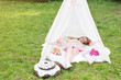 Little girl lying and playing in a tent, children's house wigwam in park. Child with big donuts and heart-shaped pillows. Summer, happy childhood concept. Cute emotions girl with pink bow.