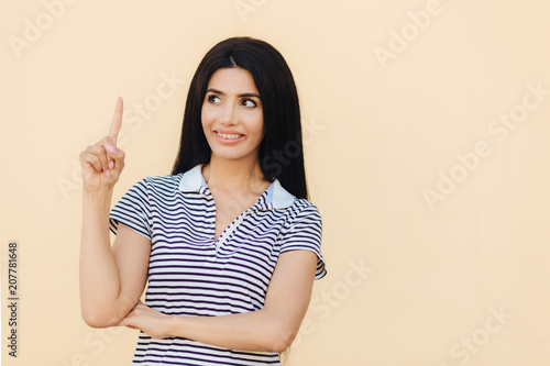 Fotografie, Obraz Glad brunette female with pleasant smile, has long dark hair, indicates with fore finger upwards, shows blank space for your advertisement or promotional background