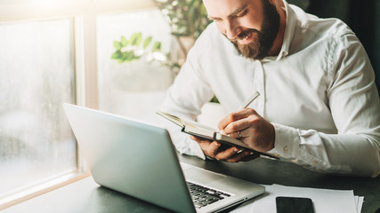 Young smiling bearded businessman in white shirt sitting at desk in front of laptop, making notes in notebook. Freelancer works remotely. Student learning online. Online traning, marketing, education.