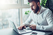 © foxyburrow - Young bearded businessman in white shirt is sitting at table and working on laptop. Freelancer works at home. Student is studyng online. Online marketing, education, blogging. Business planning.