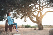 Lost in thoughts. Full length profile of charming girl is spending time outdoors with beautiful sunset and old tree in background. She is sitting on bench while looking aside thoughtfully. Copy space