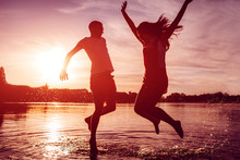 Happy Couple Jumping On Summer River Bank. Young Man And Woman Having Fun At Sunset. Guys Hanging Together