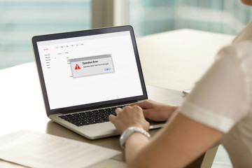 Businesswoman looks at laptop screen with Operation Error message. Entrepreneur experienced system failure during workflow. Computer system crash, software failure, malware. Rear view over shoulder.