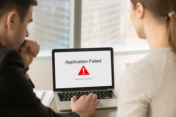 Two business people looking at laptop screen with Application Failed error message. Businessman with female client experience system failure during project demonstration. Rear view over shoulder.