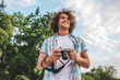 © iuricazac - Smiling attractive young tourist man with digital camera taking photos, on a nature and sky background. Handsome man curly hair exploring nature on vacation trip. Travel, people and lifestyle concept