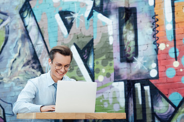 happy young businessman in earphones and eyeglasses using laptop while sitting outside