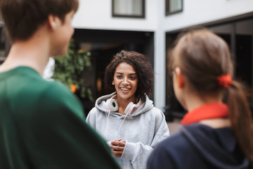 Pretty lady with dark curly hair in headphones standing and happily looking at her friends while spending time in courtyard of university