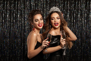 Wall Mural - Two beautiful young girls smiling and holding a glass of champagne, meeting Christmas and New Year. Clean skin and a hairstyle with bright lips. Photographed on a sparkling background.