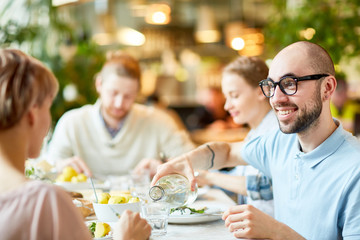 crop view of smiling bald bearded male in glasses sitting with friends and pouring water to woman