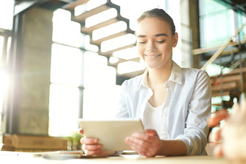 Young pretty woman sitting at table in sunlight and browsing tablet computer with smile.