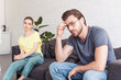 © LIGHTFIELD STUDIOS - upset young man in eyeglasses and girlfriend sitting on sofa behind at home
