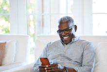 African American Senior Man Texting On A Smart Phone Sitting On The Sofa At Home