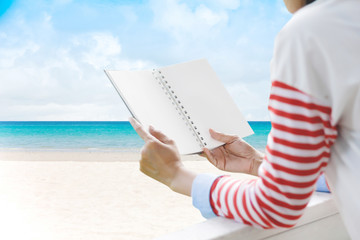 Wall Mural - Rear view of woman reading novel by the sea.Summer beach background. Sand and sea and sky