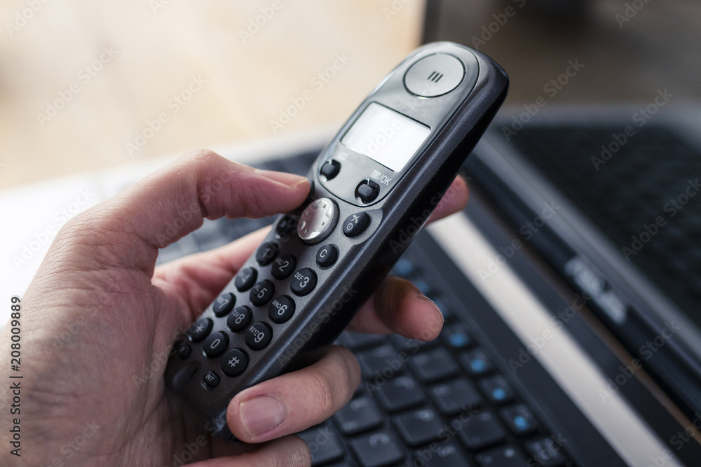 Hand Holding a Black Cordless Telephone with Laptop at Background - obrazy, fototapety, plakaty 