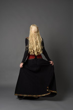 Full Length Portrait Of Pretty Blonde Lady Wearing  A Red And Black Fantasy Medieval Gown. Standing Pose On Grey Background.