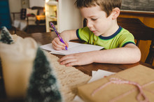 Little Boy Sitting At The Kitchen Table Signing Christmas Cards