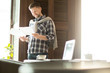 © pressmaster - Handsome man in casual clothes smiling and reading papers while standing near cafe table with laptop.