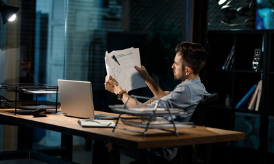 Wall Mural - Formal man sitting in office with lamp burning and reading paper documents 