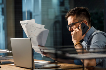 Wall Mural - Young busy man holding documents in hands and watching laptop while talking on smartphone in office alone. 