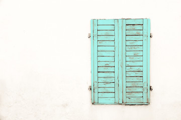 Rustic old grungy and weathered green cyan wooden closed window shutters with peeling paint on a white wall of an abandoned house.