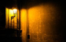 Old Lantern Illuminating A Dark Alleyway Corner Wall At Night In Prague, Czech Republic. Photo Almost Monochromatic With Brown Yellow Tones From The Lantern As Light Source Against The Dark Shadows