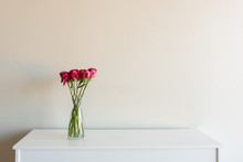 Close Up Of Variegated Red Roses In Glass Vase On White Sideboard Against Neutral Wall Background