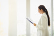 © kasipat - Business woman standing working near a window and holding clipboard at office.