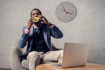 Wall Mural - mature african american man eating hamburger at table with laptop