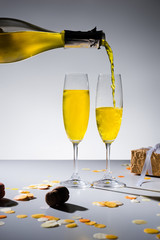 Wall Mural - close up view of pouring yellow champagne into glasses process and wrapped gift on grey backdrop