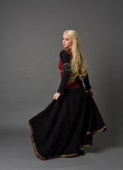 full length portrait of pretty blonde lady wearing  a red and black fantasy medieval gown. standing pose on grey background.