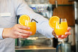 The barman gives two glasses of orange juice to the client of the hotel restaurant. The waiter transfers the order for two orange juice to the client of the hotel bar. The concept of service.
