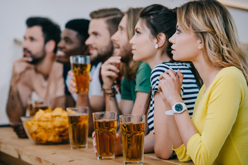  side view of group of multicultural friends drinking beer and watching soccer match at bar