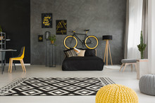 Bicycle Above Black Bed In Dark Open Space Interior With Pouf And Yellow Chair At Desk. Real Photo