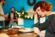 © pressmaster - Depressed red-haired woman sitting alone at table, drinking cocktail and thinking while her friends having fun in background