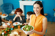 © pressmaster - Young female standing at home holding white bowl with green salad and looking at camera while eating with friends