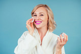 Fototapeta Sypialnia - attractive and smiling woman blonde in a cotton white bathrobe applies moisturizer cream on face in the studio on a blue background . concept of skin care and spa