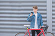 © bodnarphoto - Happy young man standing near a red bike and using a smartphone on the background of a gray wall. A student cyclist in a jeans jacket standing against the wall with a bike and smiles.