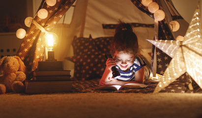 Wall Mural - happy child girl laughing and reading book in dark in tent at home