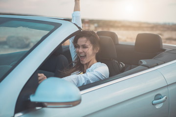 I am free. Joyful girl is driving a car in the nature. She is raising arm up with excitement and laughing