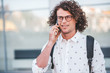 © iuricazac - People, technology, lifestyle. Portrait of stylish curly hair male in trendy spectacles standing outdoors while holding backpack and talking by his smart phone outdoor on the city street.