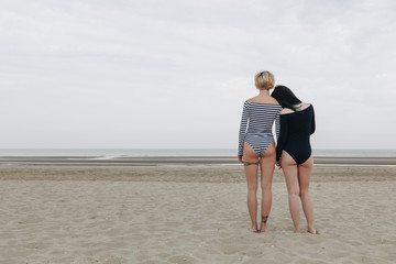 Wall Mural - rear view of attractive young women in bodysuits embracing on sandy beach on cloudy day