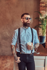 Wall Mural - Old-fashioned tattooed hipster wearing a shirt and suspenders, in a sunglasses, standing with a cup of a coffee near a desk with a laptop, looking out the window in an office with a loft interior.