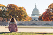 Young woman in coat sitting looking at view of United States Congress Capitol building, golden orange yellow foliage autumn fall trees on street during sunny day in Washington DC