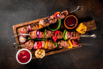 Fresh, home-cooked on the grill fire meat beef shish kebab with vegetables and spices, with barbecue sauce and ketchup, on a dark background on a wooden cutting board above copy space