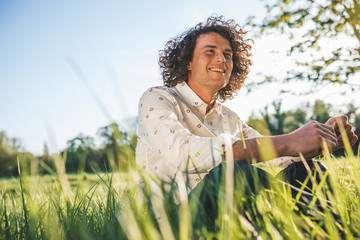 Wall Mural - Outdoor shot of happy handsome young male with curly hair smiling and looking a side, sitting on the green grass in the park, relaxing after busy day. Copy space for advertising. People, lifestyle