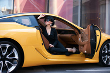 Happy Beautiful Young Woman Sitting In A Yellow Sports Car On Beautiful Sunny Summer Day. Business Woman Lady In Black Suit And Sunglasses Sitting In Luxury Supercar