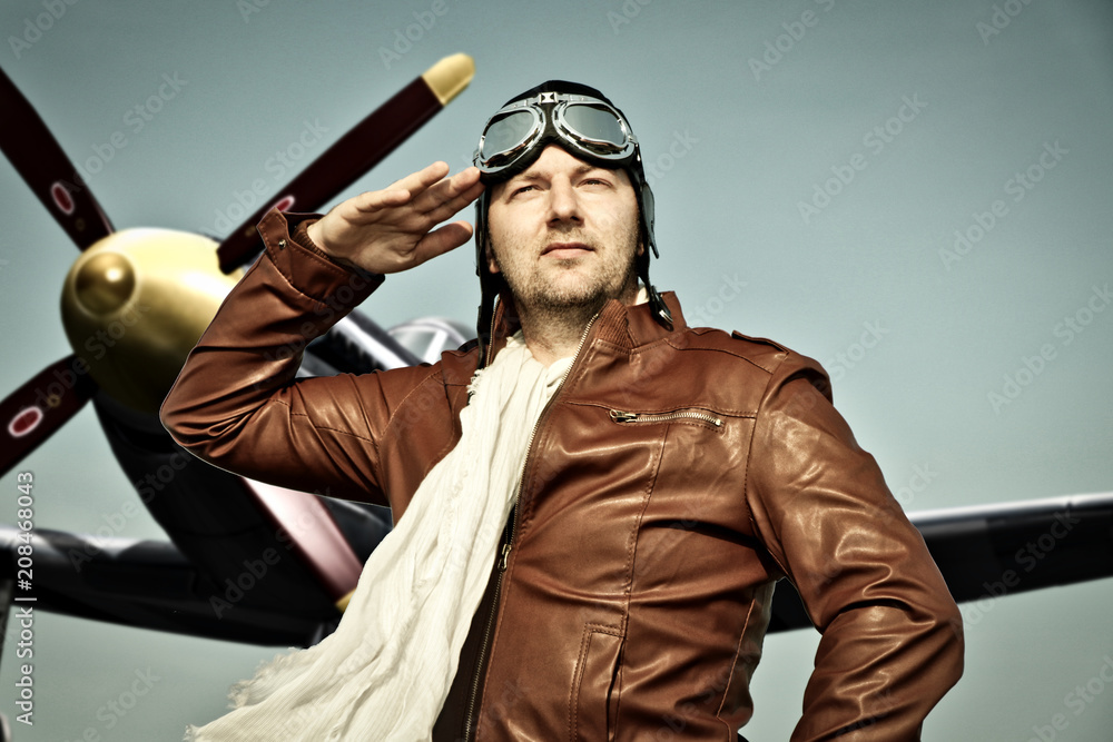 Portrait of a vintage pilot with leather cap, scarf and aviator glasses salutes in front of a historic airplane - Portrait of a man in historical pilot clothing - obrazy, fototapety, plakaty 